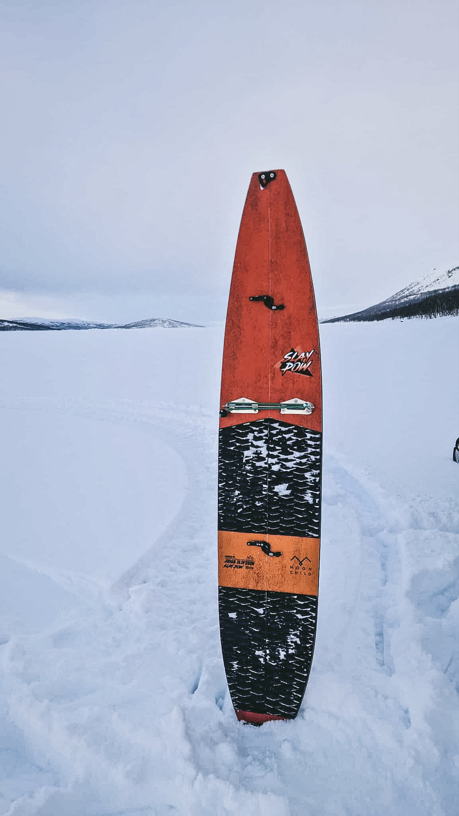 Orange Johan Olofsson's splitsurf standing vertically in the snow with a mountainous background.